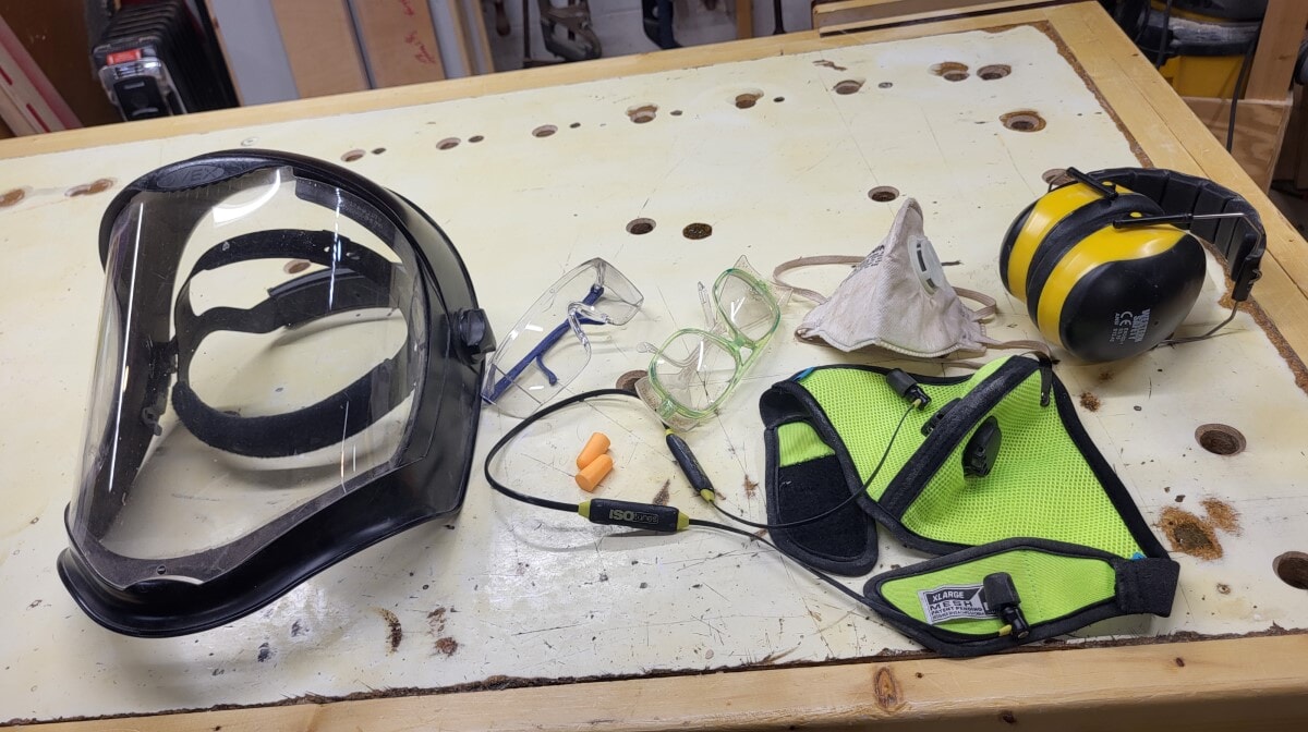 Collection of woodworking PPE including face shield, safety glasses, dust mask, ear muffs, and safety vest laid out on a workbench