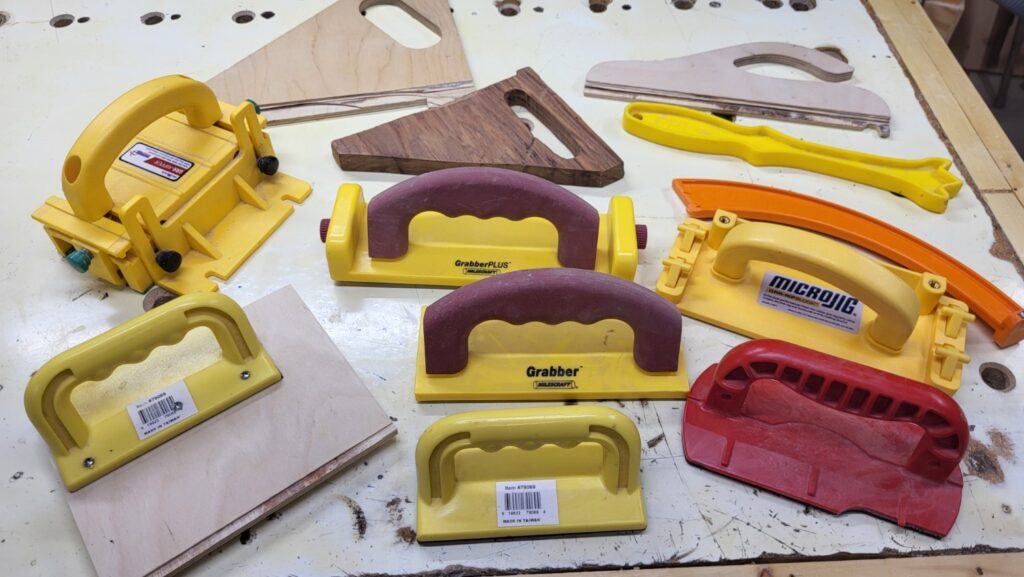 Different push blocks, push shoes, and safety tools laid out on a workbench, showing both homemade and purchased designs used to keep hands safe when cutting wood on a table saw