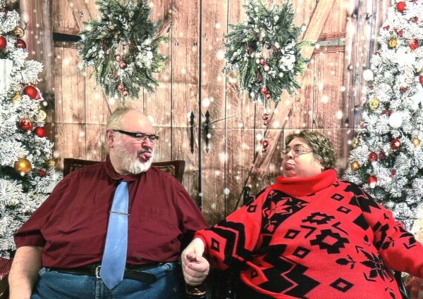 Mike and Cindy sitting together in front of a festive Christmas backdrop with wreaths and decorated trees, playfully sticking their tongues out at each other while holding hands. Their laughter and affection capture the fun and love that defined their life together.