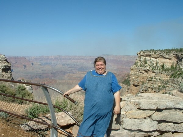 Cindy standing near the railing at the Grand Canyon, smiling as she holds on tightly while posing against the vast canyon backdrop. The bright day and endless view capture both her sense of adventure and her playful hesitation about being too close to the edge.