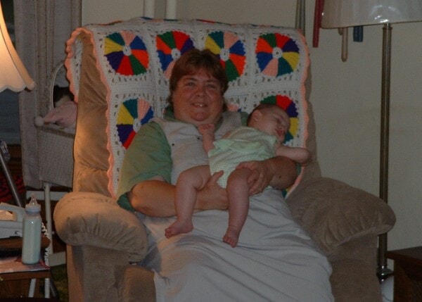 Cindy sitting in a beige recliner, holding her sleeping baby niece dressed in light green. A colorful crocheted blanket rests across the back of the chair, adding to the cozy setting. Her peaceful smile and relaxed posture reflect the quiet love and gentleness that defined her spirit.