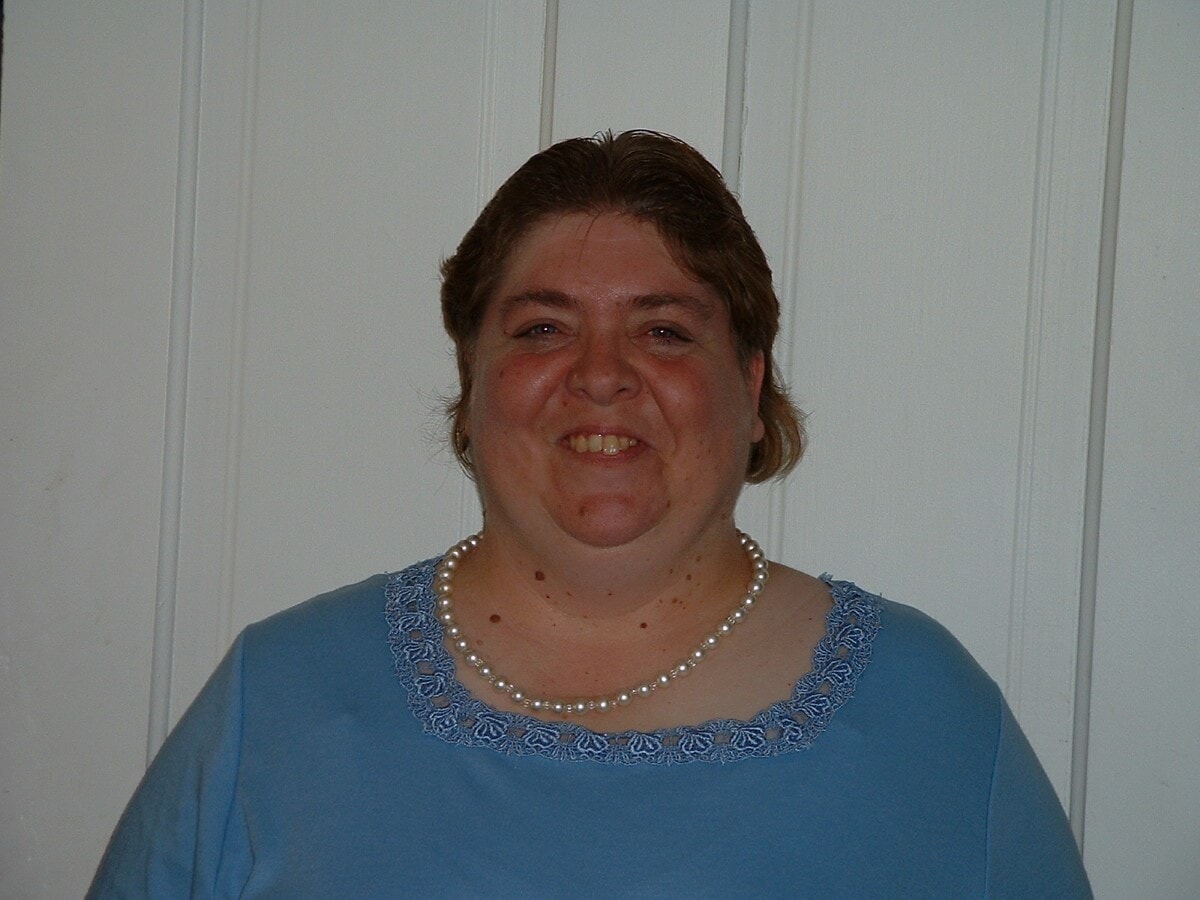 Cindy smiling in front of a white paneled wall, wearing a light blue top with lace trim and a pearl necklace. Her playful smile reflects the joy and warmth that defined her spirit.