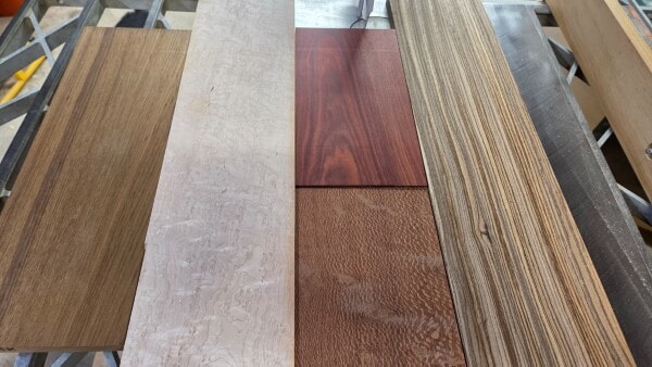 Wood samples including Teak, Birdseye Maple, Lacewood, Bloodwood, and Zebrawood arranged side by side to show contrasting colors and grain patterns.