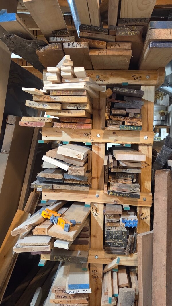 End view of a lumber rack in a woodworking shop, filled with labeled boards of different species and sizes.