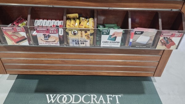 Woodcraft checkout counter bins filled with small woodworking essentials like glue, card scrapers, sanding blocks, and marking tools.