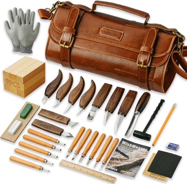 Complete wood carving kit with leather tool roll, carving knives, gouges, sharpening supplies, gloves, wood blocks, and accessories laid out on a table.